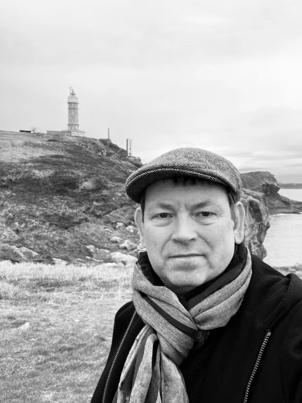 Michael Tschiggerl mit Schiebermütze und Schal an einer Steilküste, im Hintergrund ein Leuchtturm